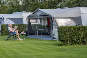 Campingzelt mit Vorzelt auf grünem Rasen, daneben ein Klappstuhl und Hecken, im Hintergrund weitere Zelte im Garten.