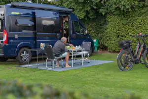 Blaues Wohnmobil mit ausgebauter Terrasse, Tisch und Stühlen, daneben Fahrräder auf dem Stellplatz im Grünen.