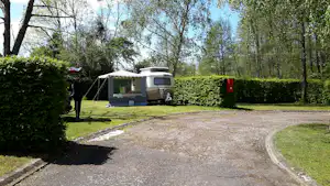 Wohnwagen auf einem Campingplatz-Stellplatz, umgeben von Hecken und Bäumen, mit gepflastertem Weg und Rasenfläche.