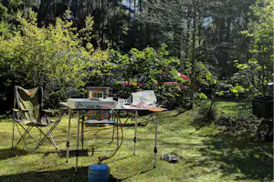 Camping-Tisch mit Grill und Geschirr, daneben Klappstühle im Grünen, im Hintergrund Bäume und ein rotes Auto.