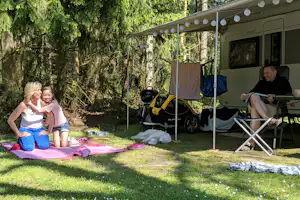 Wohnmobil mit Vorzelt, Campingstühlen und Decke auf dem Rasen, im Hintergrund hohe Bäume.