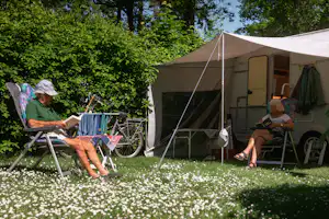 Wohnwagen mit Markise, davor Campingstühle und Fahrrad im Grünen unter Bäumen.