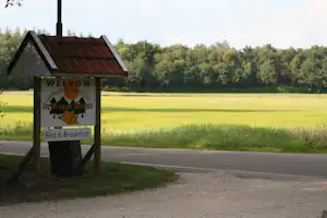 Willkommensschild am Campingplatz am Straßenrand, dahinter Wiese und Bäume, im Hintergrund ein Feldweg.