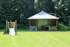 Campingzelt mit Terrasse auf dem Rasen, daneben Spielgerüst und Bälle im Garten, umgeben von Bäumen.