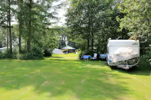 Wohnwagen auf einem grünen Campingplatz mit Rasen, Bäumen und einem weiteren Zelt im Hintergrund.