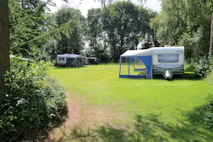 Wohnwagen und Zelt auf einer grünen Wiese zwischen Büschen und Bäumen, mit Schotterweg im Vordergrund.