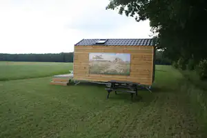 Holzhütte auf Stelzen auf einer Wiese, mit großem Bild an der Außenwand und Bäumen am Rand.