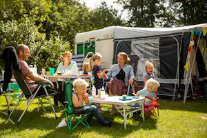 Campingfamilie sitzt am Tisch vor einem Zelt, Kinder essen und trinken auf dem Campingplatz im Grünen.