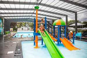 Kinder rutschen auf einer grünen Rutsche im überdachten Poolbereich, mit Becken und Geländer im Hintergrund.