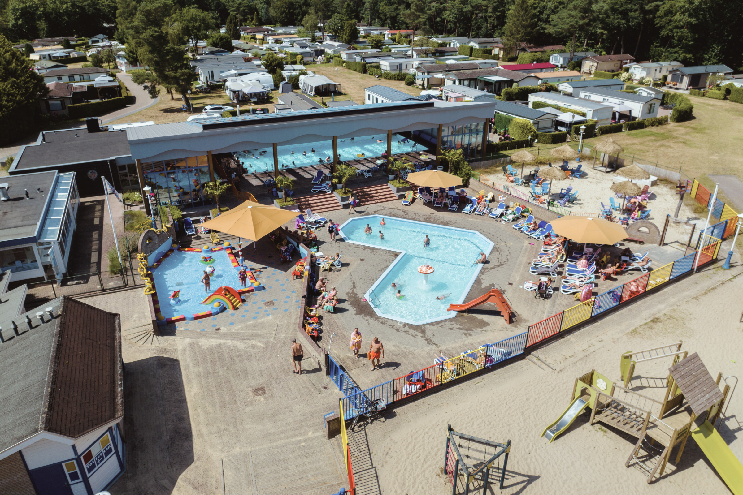 Luftaufnahme von zwei Schwimmbecken auf dem Campingplatz, umgeben von Liegen, Sonnenschirmen und Wohnmobilen.