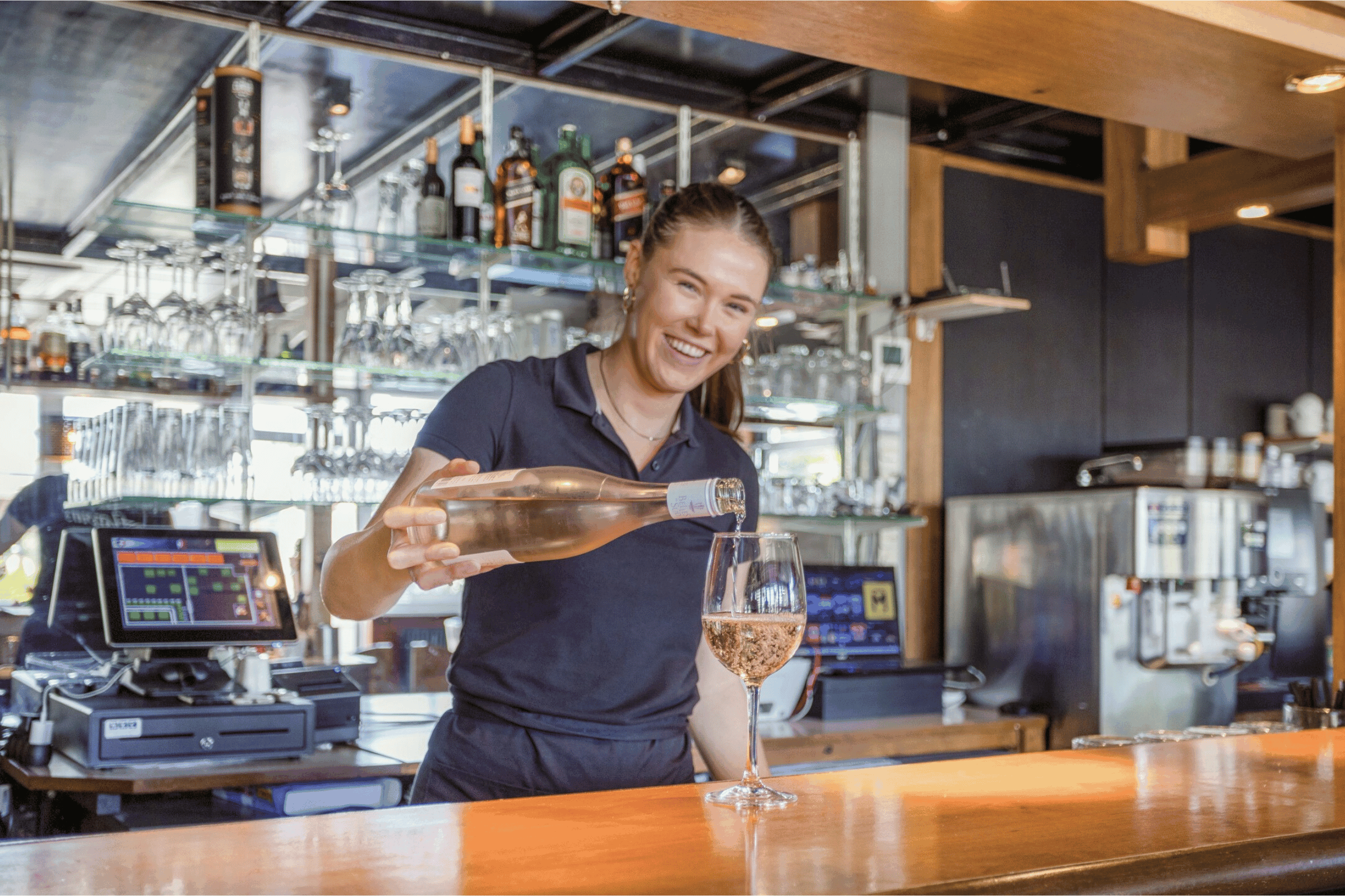 Barpersonal schenkt ein Glas Wein an der Theke in einer Camping-Bar aus, im Hintergrund Gläser und Getränkeauswahl.