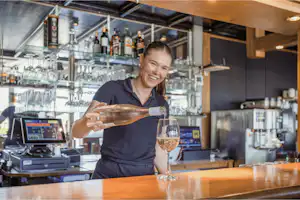Barpersonal schenkt ein Glas Wein an der Theke in einer Camping-Bar aus, im Hintergrund Gläser und Getränkeauswahl.