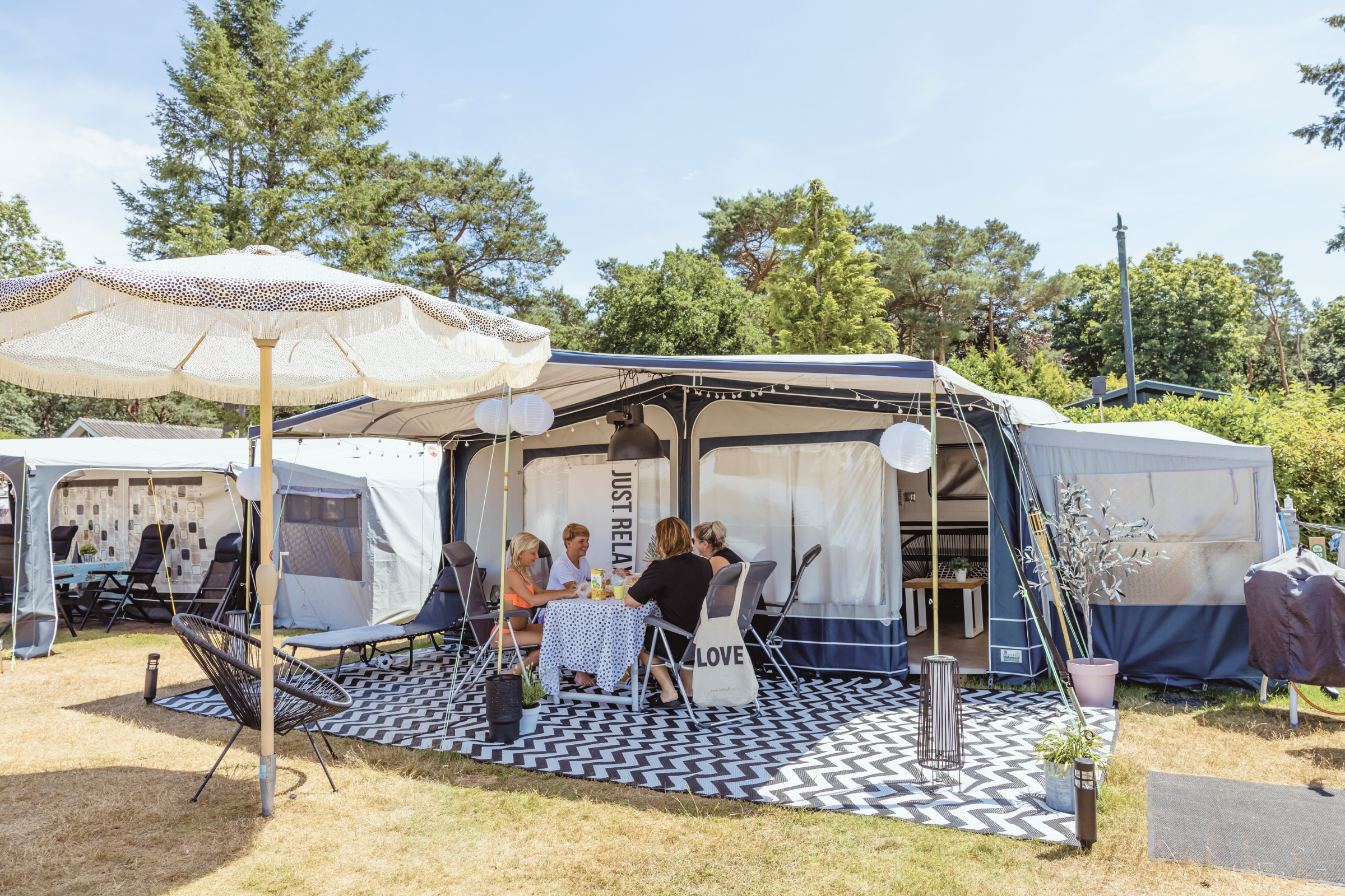 Campingzelte nebeneinander mit Sonnenschirm, gepolsterten Stühlen und Tisch auf einem gepflasterten Stellplatz, im Hintergrund Bäume.
