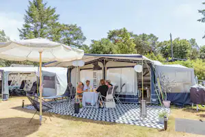 Campingzelte nebeneinander mit Sonnenschirm, gepolsterten Stühlen und Tisch auf einem gepflasterten Stellplatz, im Hintergrund Bäume.