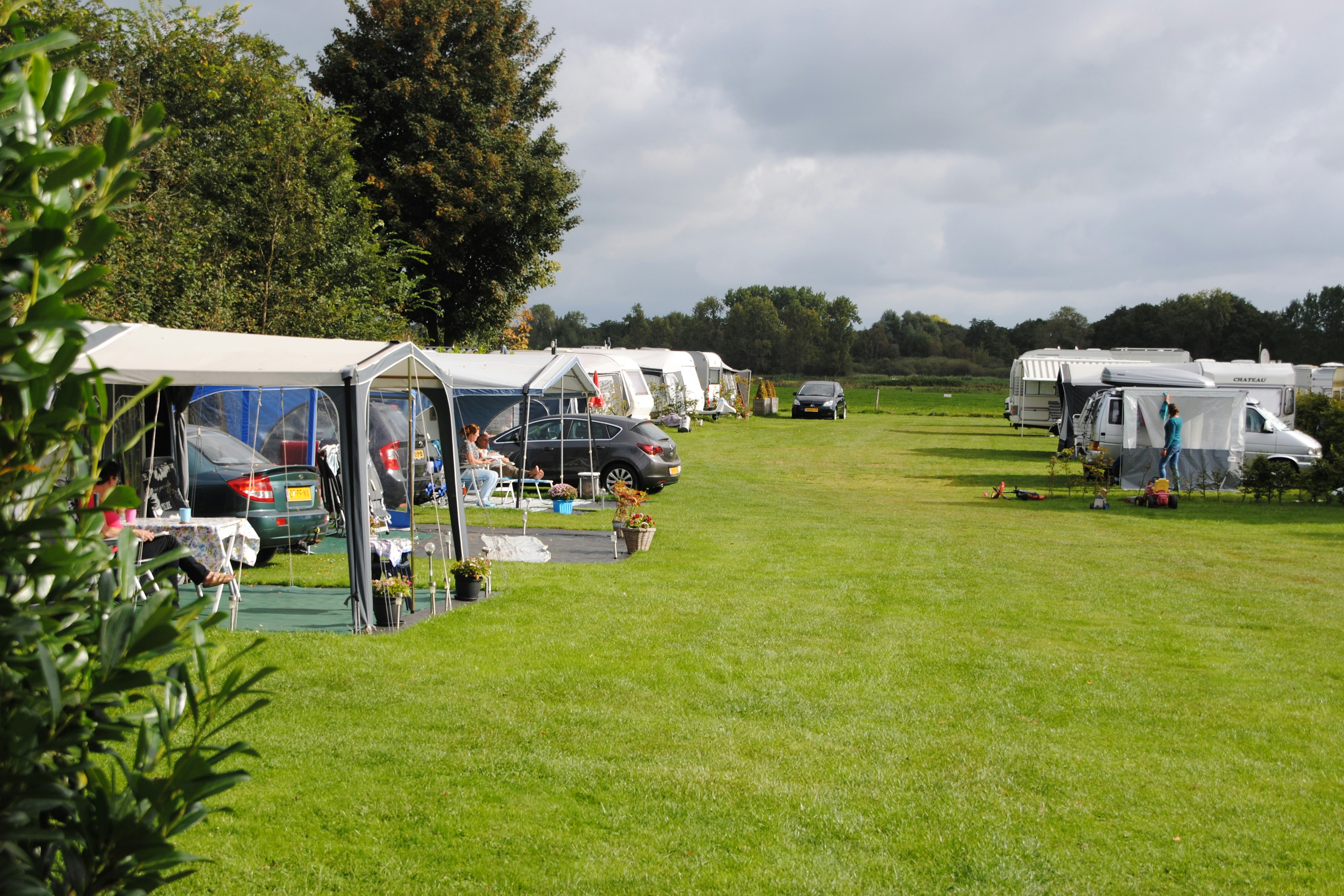 Campingplatz mit Stellplätzen auf grünem Rasen, Wohnwagen und Autos, daneben ein offenes Zelt mit Sitzbereich.