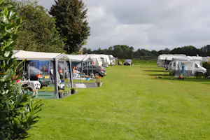 Campingplatz mit Stellplätzen auf grünem Rasen, Wohnwagen und Autos, daneben ein offenes Zelt mit Sitzbereich.