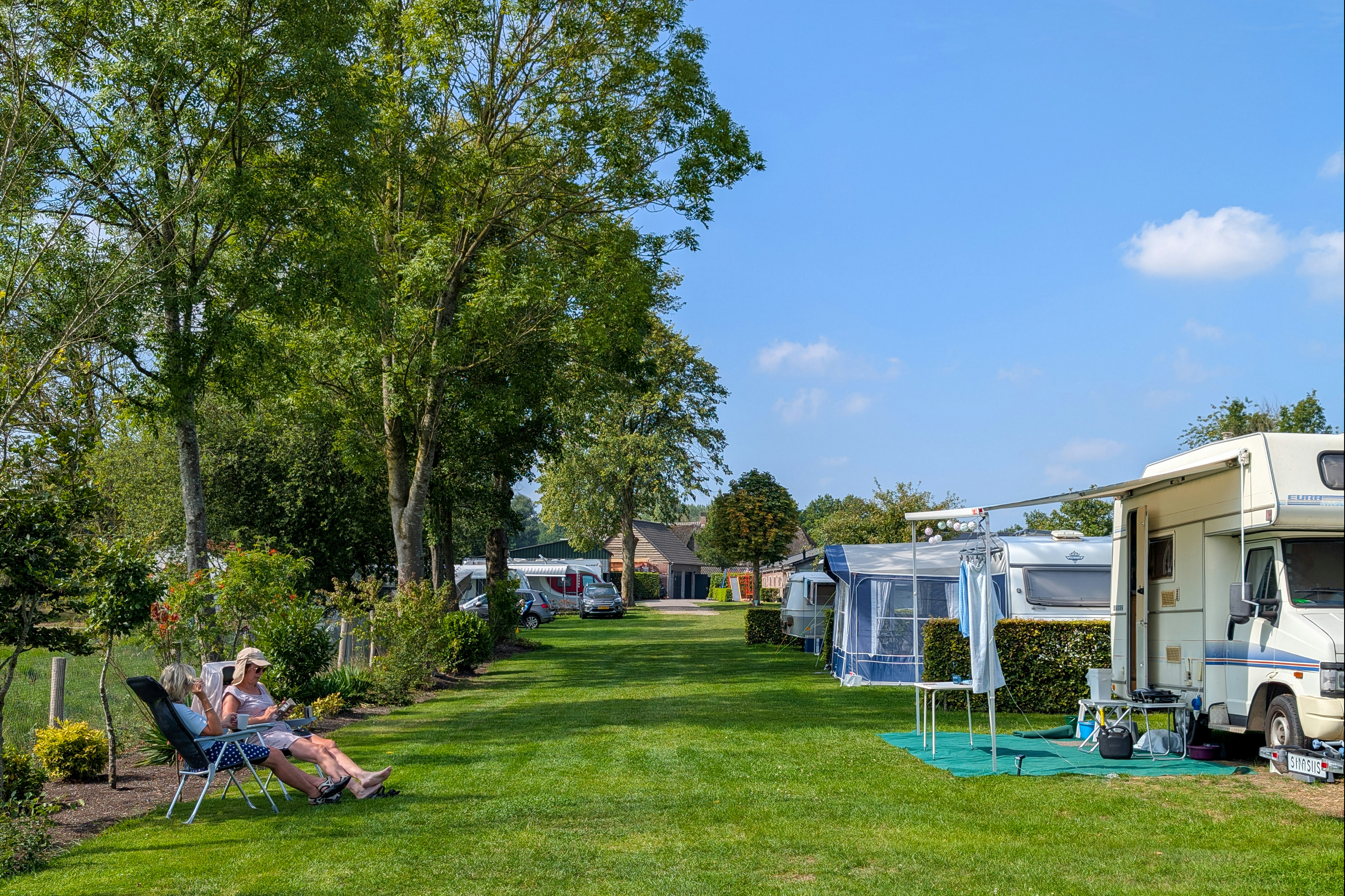 Campingplatz mit Wohnmobil und Vorzelt, gepflegtem Rasen und Sitzgelegenheiten; im Hintergrund weitere Stellplätze und Bäume.