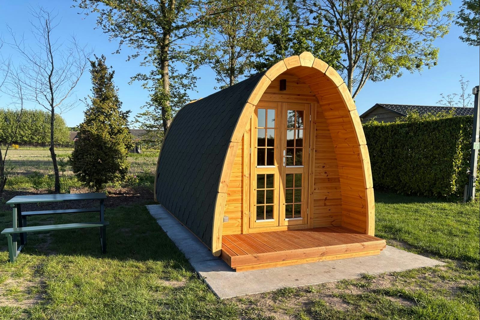 Holz-Blockhütte mit halbrunder Vorderseite und Tür, auf Betonpodest im Garten, umgeben von Bäumen und Hecke.