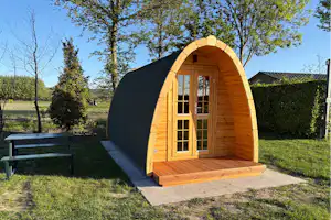 Holz-Blockhütte mit halbrunder Vorderseite und Tür, auf Betonpodest im Garten, umgeben von Bäumen und Hecke.