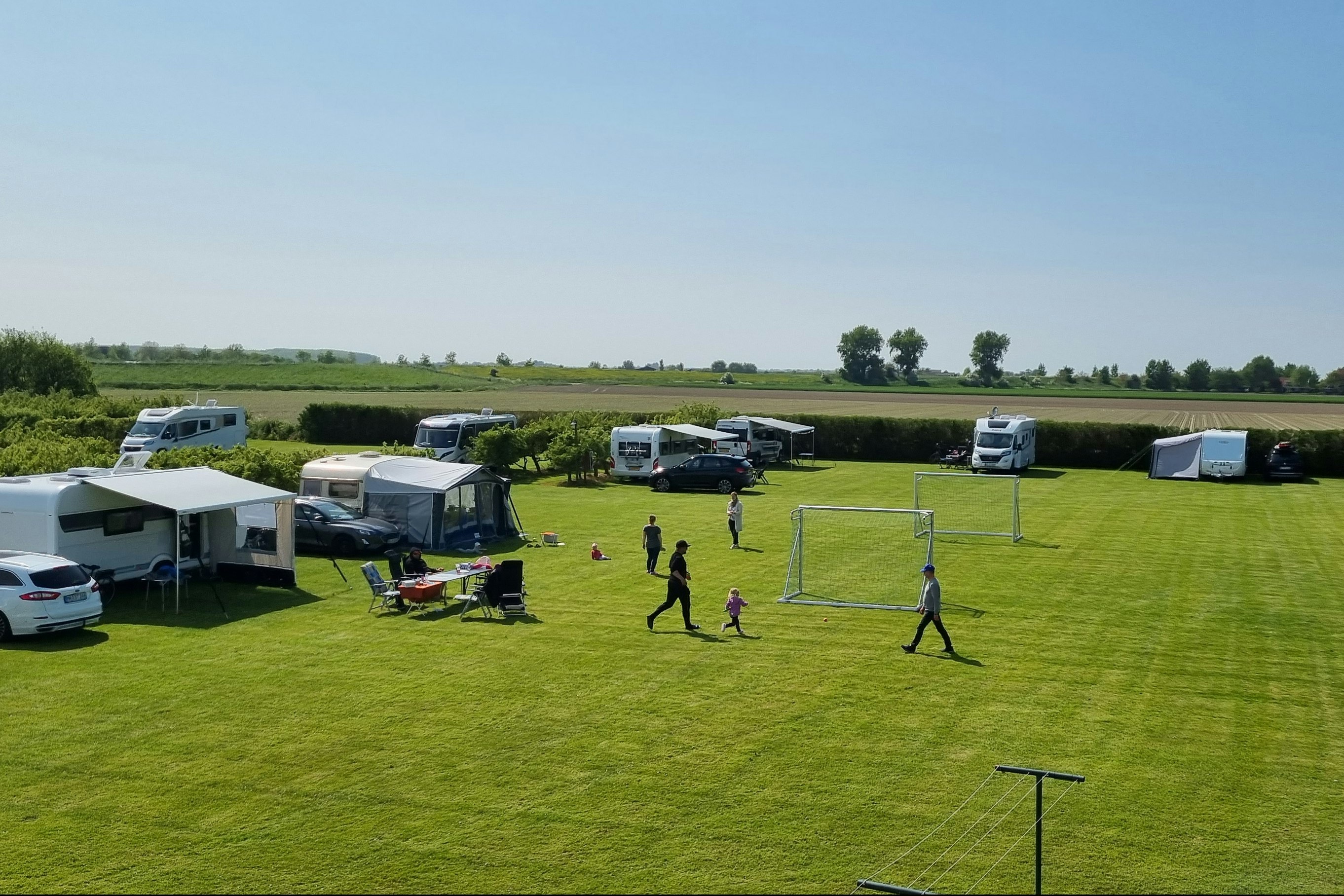 Campingplatz mit Wohnmobilen und Zelten auf einer großen Rasenfläche, daneben ein Volleyballfeld und ein Weg zwischen den Stellplätzen.
