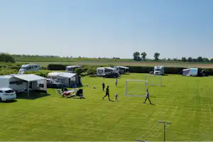 Campingplatz mit Wohnmobilen und Zelten auf einer großen Rasenfläche, daneben ein Volleyballfeld und ein Weg zwischen den Stellplätzen.