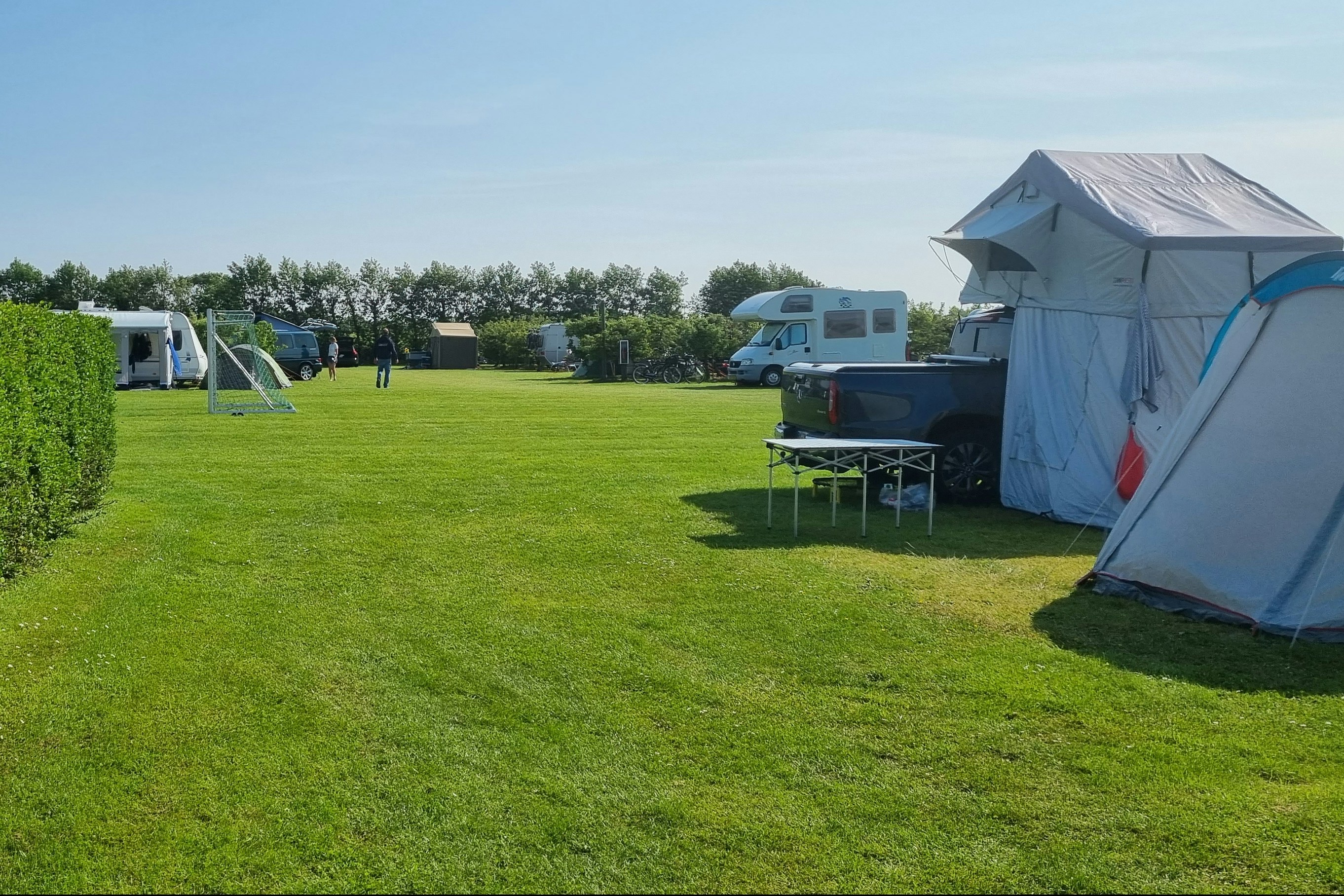 Zelt auf einem grünen Campingplatz, daneben Tisch und Stühle, Hecken und weitere Zelte im Hintergrund.