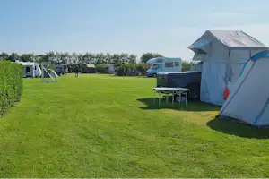 Zelt auf einem grünen Campingplatz, daneben Tisch und Stühle, Hecken und weitere Zelte im Hintergrund.