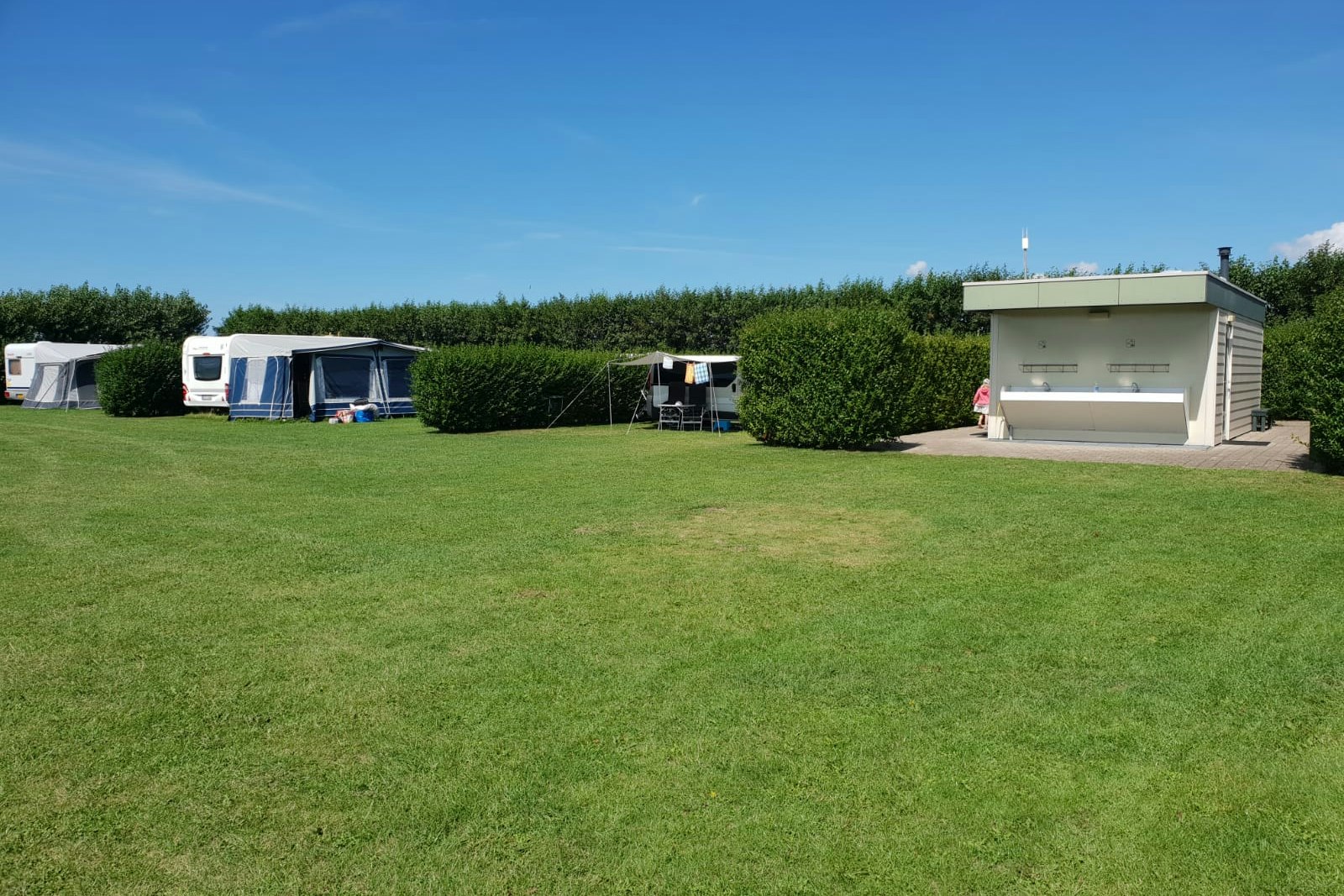 Campingplatz mit gepflegtem Rasen, mehreren Zelten und Wohnwagen sowie einem kleinen Gebäude am Rand, Hecken und Himmel im Hintergrund.