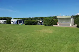 Campingplatz mit gepflegtem Rasen, mehreren Zelten und Wohnwagen sowie einem kleinen Gebäude am Rand, Hecken und Himmel im Hintergrund.