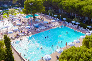 Großes Camping-Schwimmbecken mit vielen Liegen und blauen Sonnenschirmen, umgeben von Bäumen und Ferienhäusern.