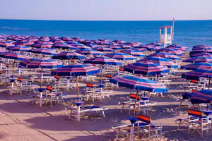 Reihe von Strandliegen mit blau-rot gestreiften Sonnenschirmen am Sandstrand, im Hintergrund das Meer und ein Rettungsturm.