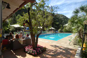 Blick auf den Camping-Pool mit blauem Wasser, Liegen und Sonnenschirmen, umgeben von Bäumen und Blumenbeeten.