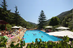 Großes Camping-Schwimmbecken mit Sonnenschirmen, Liegen und mehreren Personen im Wasser, umgeben von Bäumen und Hügeln.