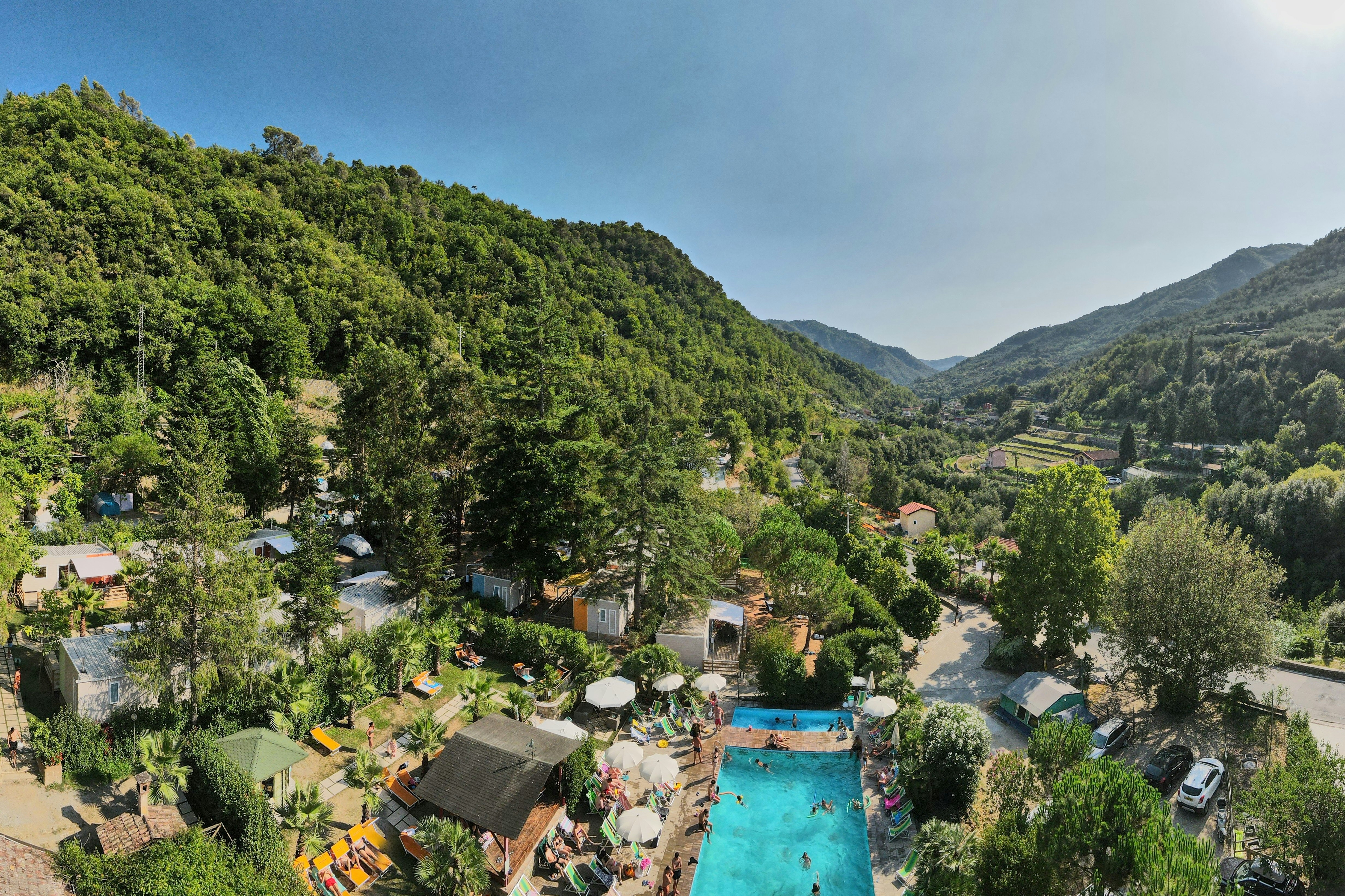 Campingplatz mit Pool inmitten von grünen Hügeln, Zelt- und Wohnwagenstellplätzen sowie Wegen zwischen Bäumen und kleinen Gebäuden.