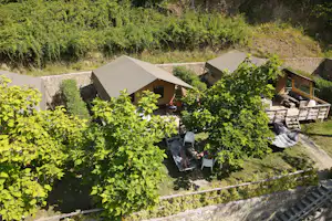 Zelt und Holzhütten auf einem Campingplatz am Hang, umgeben von Weinreben, mit Sitzbereich und Wegen zwischen den Unterkünften.