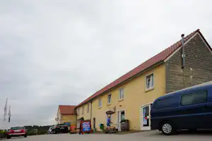 Campinggebäude mit gelber Fassade, rotem Ziegeldach und Eingang; vor dem Haus stehen Autos und ein Schild.