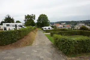 Wohnwagen auf Campingstellplätzen, Hecken und gepflasterter Weg zwischen den Parzellen, im Hintergrund Häuser und Bäume.