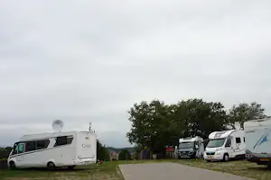 Wohnmobile auf einem Stellplatz am Rand von Grünflächen, mit gepflastertem Weg und weiteren Fahrzeugen im Hintergrund.