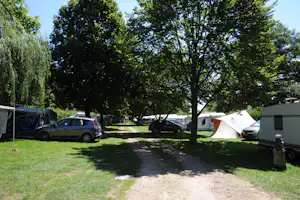 Campingplatz mit Zelt und Wohnwagen, daneben geparkte Autos auf einer Rasenfläche unter Bäumen.