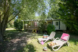Wohnwagen mit kleiner Terrasse und Holzgeländer im Grünen, daneben Gartenliegen und Spielzeug auf dem Rasen.
