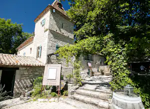 Steinhaus mit Schindeldach und Eingang am Campingplatz, gepflasterter Weg, Schild und Fassfass im Vordergrund, umgeben von Bäumen.