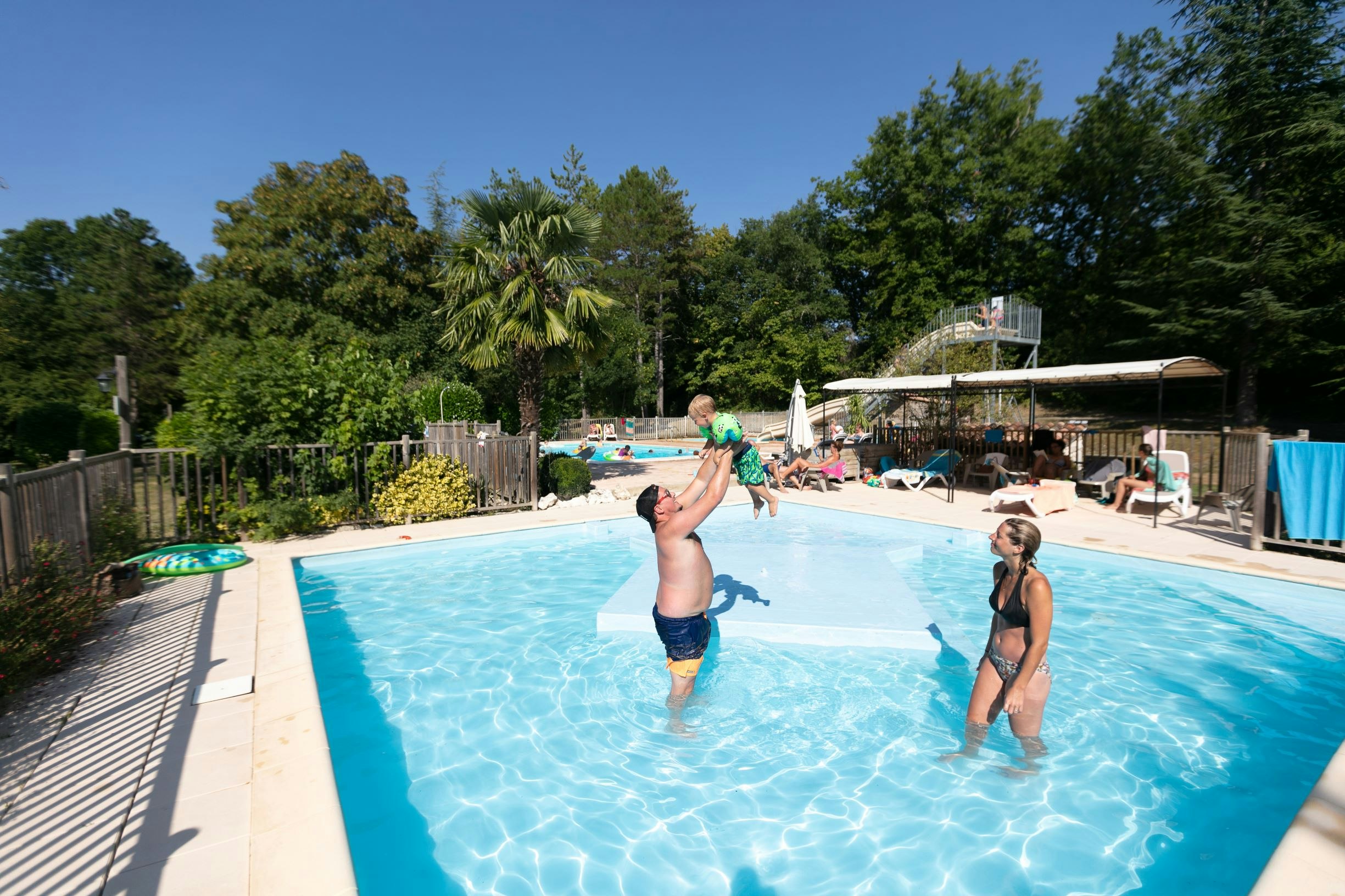 Kinder im Schwimmbecken eines Campingplatzes, umgeben von Liegen, Sonnenschirmen und Bäumen im Hintergrund.