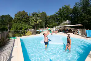 Kinder im Schwimmbecken eines Campingplatzes, umgeben von Liegen, Sonnenschirmen und Bäumen im Hintergrund.