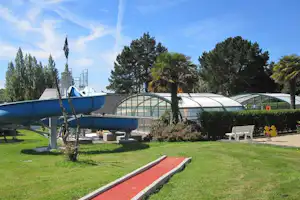 Wasserrutsche und Pool auf dem Campingplatz, mit Rasenfläche, Palmen und Hecken sowie weißen Liegestühlen im Hintergrund.
