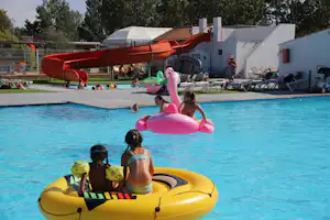 Kinder im Planschbecken und auf Schwimmreifen im Pool, daneben Liegen und Sonnenschirme vor weißen Wohnwagen am Campingplatz.
