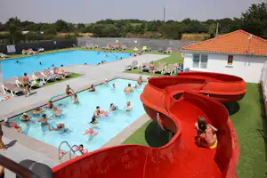 Roter Wasserrutschenturm am Pool, daneben Liegen und Kinder im Wasser vor einem gepflegten Campingbereich.