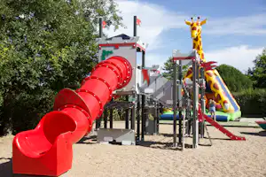 Rote Rutsche und Spielgeräte auf einem Camping-Spielplatz, mit Sandboden, Bäumen und blauem Himmel im Hintergrund.