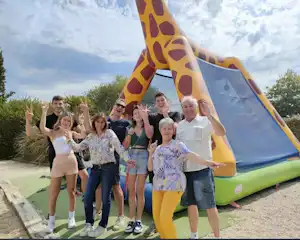 Familie vor einem aufblasbaren Spielzelt am Campingplatz, mit Kindern und Erwachsenen, im Hintergrund Bäume und Kiesweg.