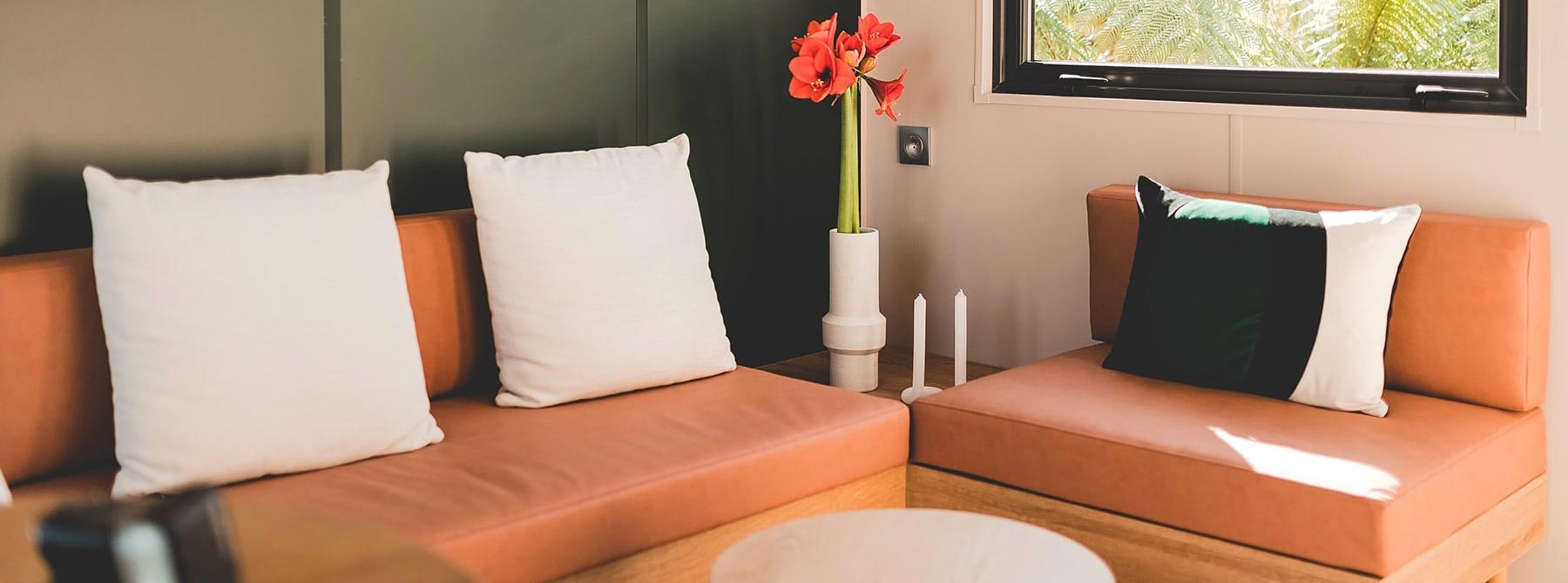 Orangefarbene Sitzbank mit weißen Kissen und schwarzem Kissen, daneben Tisch mit Vase und Fensterblick ins Grüne.