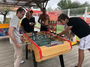 Tischkicker auf einer Holzterrasse im Campingbereich, mehrere Männer spielen unter einem überdachten Bereich.
